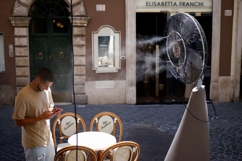 Seorang pria bernama David memeriksa ponselnya saat dia berdiri di dekat kipas angin untuk mendinginkan diri, selama gelombang panas di seluruh Italia, di Roma, 14 Juli 2023. (REUTERS/Guglielmo Mangiapane)