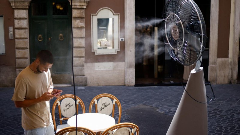 Seorang pria bernama David memeriksa ponselnya saat dia berdiri di dekat kipas angin untuk mendinginkan diri, selama gelombang panas di seluruh Italia, di Roma, 14 Juli 2023. (REUTERS/Guglielmo Mangiapane)