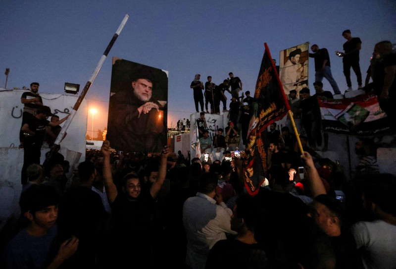 A protester holds up a portrait of Iraqi Shi'ite cleric Moqtada al-Sadr as smoke rises from the Swedish embassy building during a protest near the embassy, hours after it was stormed and set on fire ahead of an expected Koran burning in Stockholm, in Baghdad, Iraq, July 20, 2023. REUTERS/Ahmed Saad