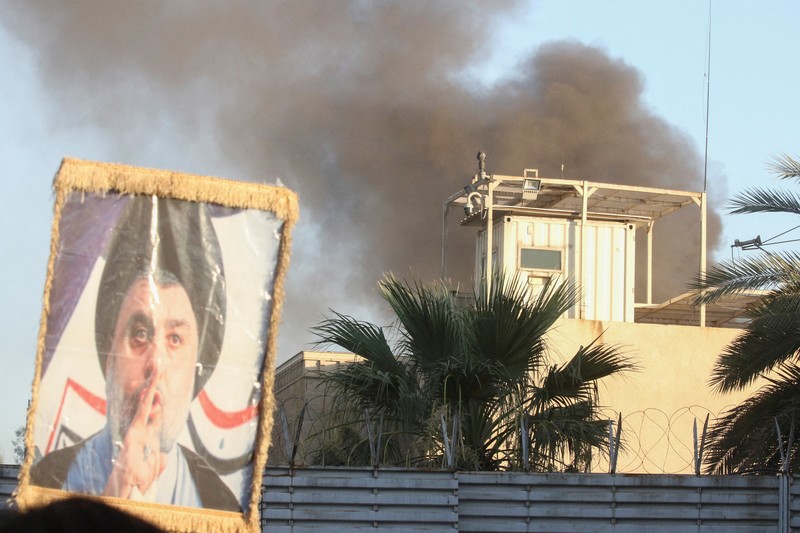 A protester holds up a portrait of Iraqi Shi'ite cleric Moqtada al-Sadr as smoke rises from the Swedish embassy building during a protest near the embassy, hours after it was stormed and set on fire ahead of an expected Koran burning in Stockholm, in Baghdad, Iraq, July 20, 2023. REUTERS/Ahmed Saad