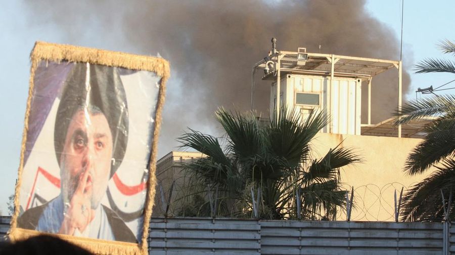 A protester holds up a portrait of Iraqi Shi'ite cleric Moqtada al-Sadr as smoke rises from the Swedish embassy building during a protest near the embassy, hours after it was stormed and set on fire ahead of an expected Koran burning in Stockholm, in Baghdad, Iraq, July 20, 2023. REUTERS/Ahmed Saad