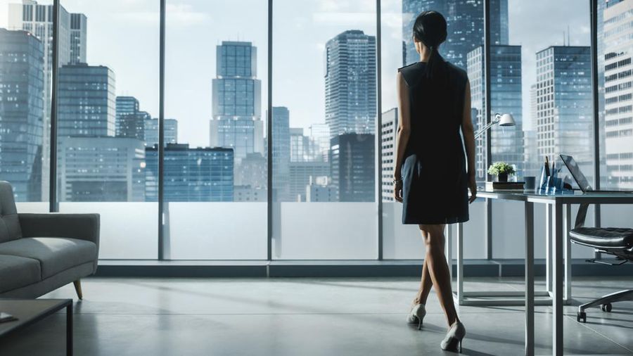 Successful Businesswoman in Stylish Dress Looking out of the Window at Big City in Downtown Area. Confident Female CEO Working on Financial Projects. Manager at Work Planning Marketing Campaign.