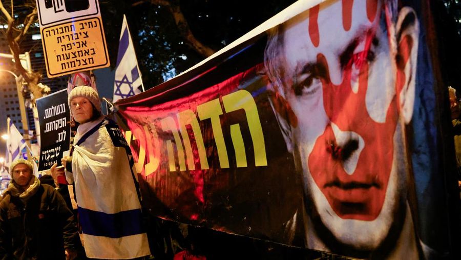 Seorang pengunjuk rasa berdiri di samping spanduk bergambar Perdana Menteri Israel Benjamin Netanyahu saat ia menghadiri rapat umum yang menuntut gencatan senjata segera di Gaza, di tengah konflik yang sedang berlangsung antara Israel dan kelompok Islam Palestina Hamas, di Tel Aviv, Israel, 27 Januari. 2024. (REUTERS/Tyrone Siu)