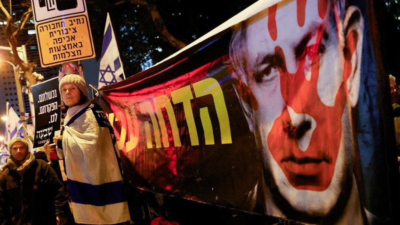 Seorang pengunjuk rasa berdiri di samping spanduk bergambar Perdana Menteri Israel Benjamin Netanyahu saat ia menghadiri rapat umum yang menuntut gencatan senjata segera di Gaza, di tengah konflik yang sedang berlangsung antara Israel dan kelompok Islam Palestina Hamas, di Tel Aviv, Israel, 27 Januari. 2024. (REUTERS/Tyrone Siu)