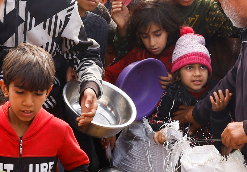 Anak-anak Palestina yang terlantar menunggu untuk menerima makanan gratis di tenda kamp, ​​di tengah kekurangan makanan, saat konflik antara Israel dan Hamas berlanjut, di Rafah di selatan Jalur Gaza, 27 Februari 2024. (REUTERS/Ibraheem Abu Mustafa)