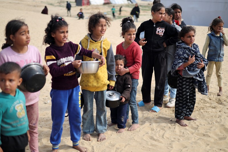 Anak-anak Palestina yang terlantar menunggu untuk menerima makanan gratis di tenda kamp, ​​di tengah kekurangan makanan, saat konflik antara Israel dan Hamas berlanjut, di Rafah di selatan Jalur Gaza, 27 Februari 2024. (REUTERS/Ibraheem Abu Mustafa)