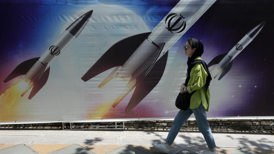 A woman walks past a banner showing missiles being launched, in northern Tehran, Iran, Friday, April 19, 2024. Iran fired air defenses at a major air base and a nuclear site near the central city of Isfahan after spotting drones early Friday morning, raising fears of a possible Israeli strike in retaliation for Tehran's unprecedented drone-and-missile assault on the country. On the missiles, a decorative sign reads: 