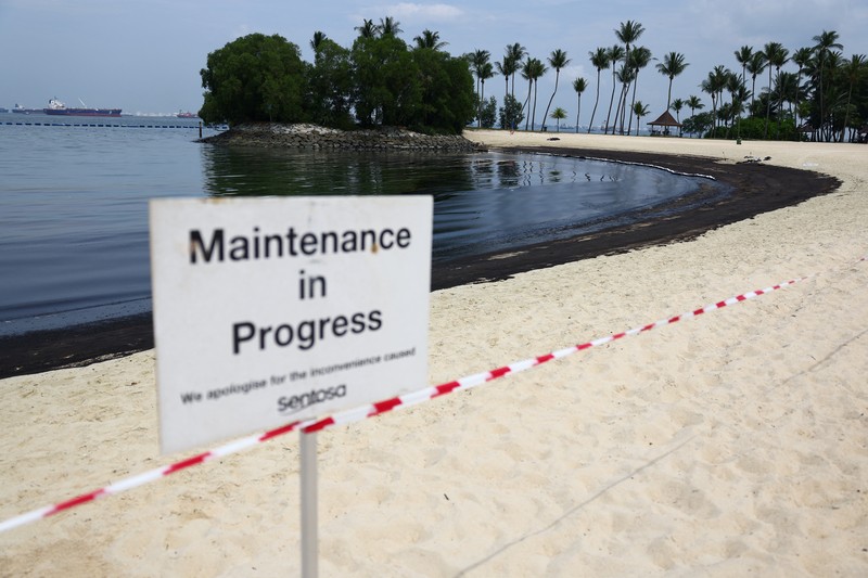 Seorang pria duduk di dekat papan tanda dan pita penjagaan dipasang menyusul tumpahan minyak di Pantai Tanjong di Sentosa, Singapura 15 Juni 2024. (REUTERS/Edgar Su)