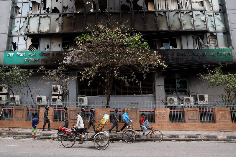 Petugas keamanan berjalan melewati kendaraan organisasi milik pemerintah yang rusak, yang dibakar oleh massa selama bentrokan setelah kekerasan meletus menyusul protes mahasiswa terhadap kuota pekerjaan pemerintah, di Dhaka, Bangladesh, 22 Juli 2024. (REUTERS/Mohammad Ponir Hossain)