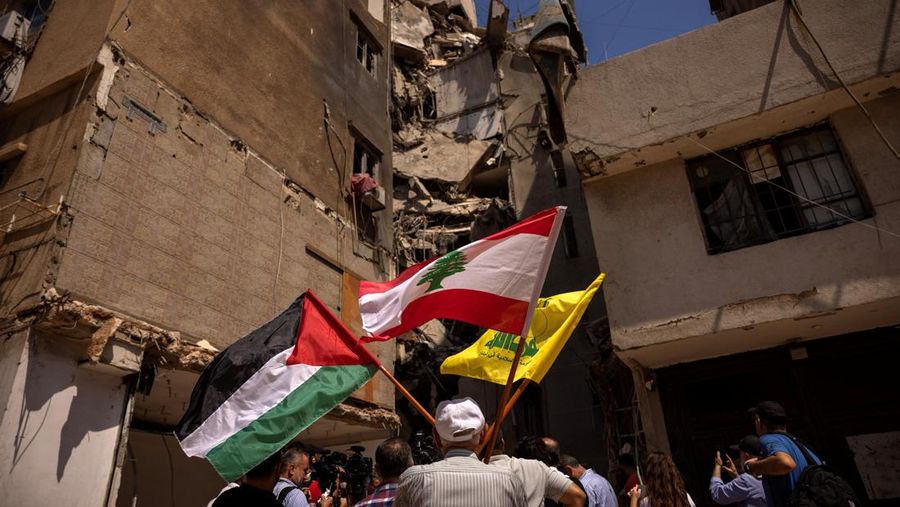 Seorang pria memegang bendera Palestina, Lebanon, dan Hizbullah di samping lokasi yang rusak di mana komandan tinggi Hizbullah Fuad Shukr terbunuh setelah serangan Israel pada tanggal 30 Juli, saat anggota Komite Kesehatan Parlemen Lebanon mengunjungi daerah tersebut, di pinggiran selatan Beirut, Lebanon, 5 Agustus 2024. (REUTERS/Alkis Konstantinidis)