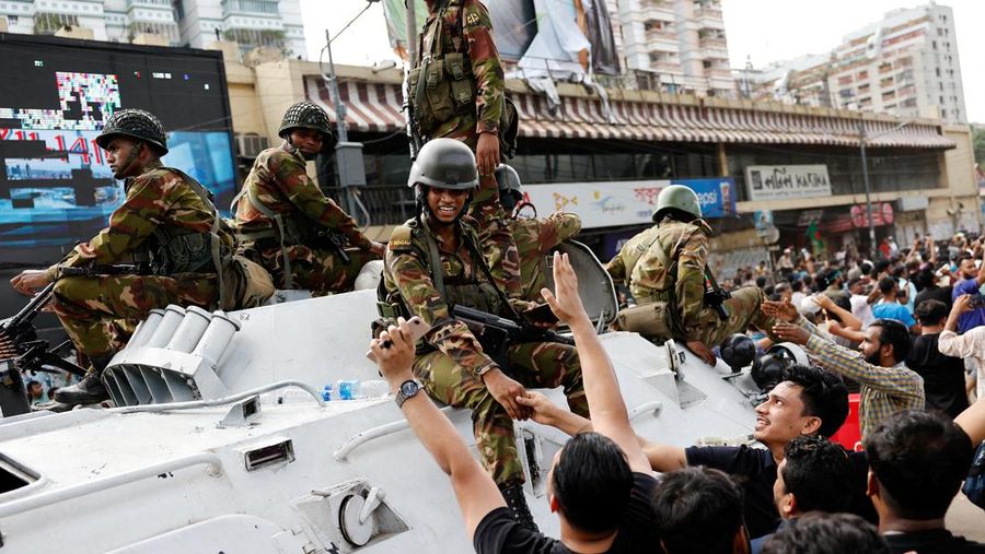 Para pengunjuk rasa antipemerintah berbaris menuju istana Perdana Menteri Sheikh Hasina sementara personel militer (tengah) berjaga di kawasan Shahbag, dekat Universitas Dhaka di Dhaka pada 5 Agustus 2024. Protes di Bangladesh yang dimulai sebagai demonstrasi yang dipimpin mahasiswa terhadap aturan perekrutan pemerintah pada bulan Juli mencapai puncaknya pada tanggal 5 Agustus, saat perdana menteri melarikan diri dan militer mengumumkan akan membentuk pemerintahan sementara. (Photo by Munir UZ ZAMAN / AFP)