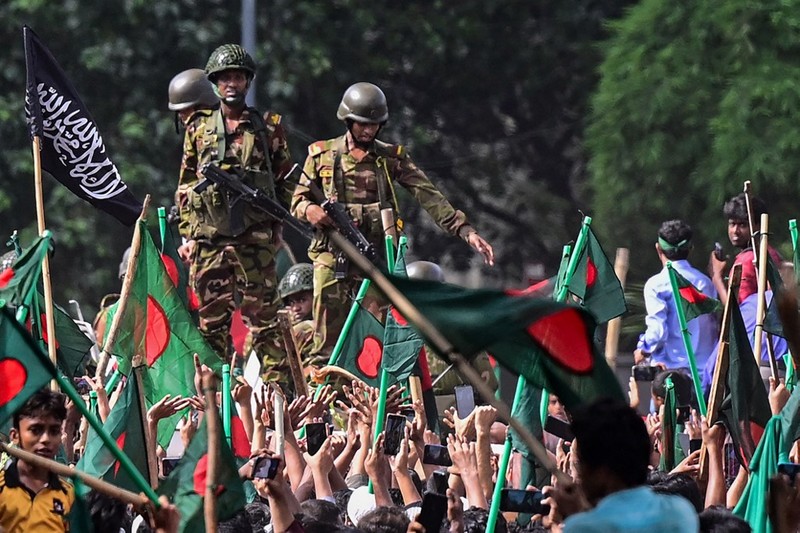 Para pengunjuk rasa antipemerintah berbaris menuju istana Perdana Menteri Sheikh Hasina sementara personel militer (tengah) berjaga di kawasan Shahbag, dekat Universitas Dhaka di Dhaka pada 5 Agustus 2024. Para pengunjuk rasa antipemerintah berbaris menuju istana Perdana Menteri Sheikh Hasina sementara personel militer (tengah) berjaga di kawasan Shahbag, dekat Universitas Dhaka di Dhaka pada 5 Agustus 2024. Protes di Bangladesh yang dimulai sebagai demonstrasi yang dipimpin mahasiswa terhadap aturan perekrutan pemerintah pada bulan Juli mencapai puncaknya pada tanggal 5 Agustus, saat perdana menteri melarikan diri dan militer mengumumkan akan membentuk pemerintahan sementara. (Photo by Munir UZ ZAMAN / AFP)