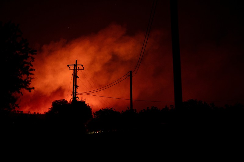 Penduduk setempat berdiri di samping mobil saat asap mengepul dari kebakaran hutan yang terjadi di desa Varnavas, dekat Athena, Yunani, 11 Agustus 2024. (REUTERS/Hilary Swift)