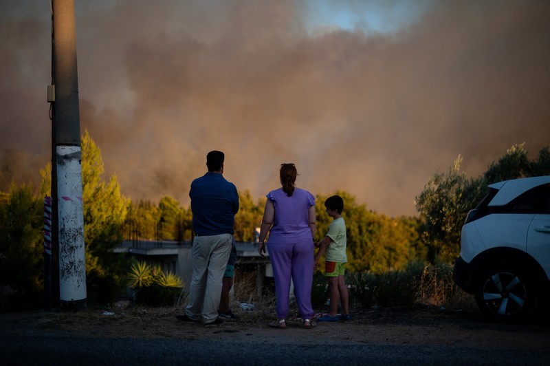 Penduduk setempat berdiri di samping mobil saat asap mengepul dari kebakaran hutan yang terjadi di desa Varnavas, dekat Athena, Yunani, 11 Agustus 2024. (REUTERS/Hilary Swift)