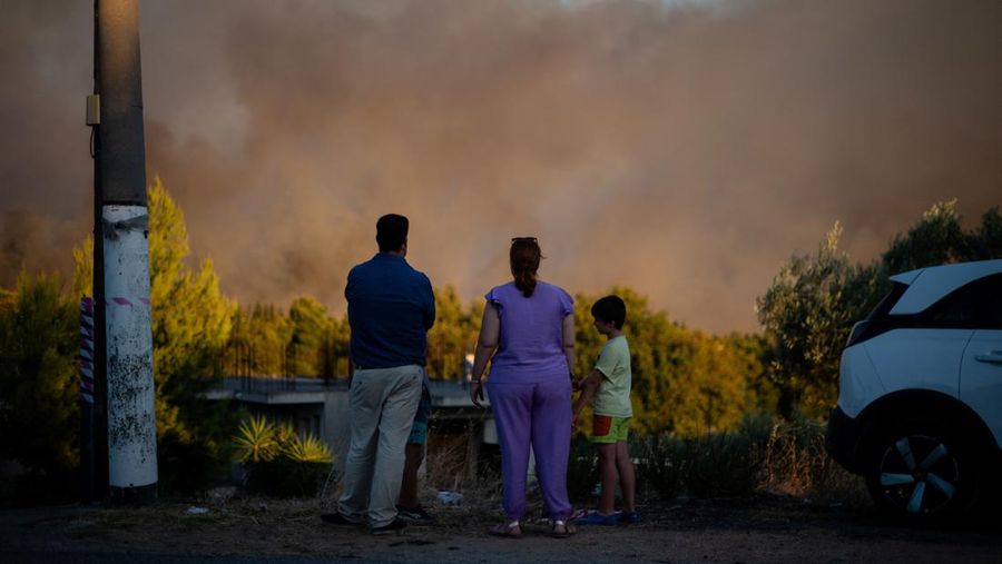 Penduduk setempat berdiri di samping mobil saat asap mengepul dari kebakaran hutan yang terjadi di desa Varnavas, dekat Athena, Yunani, 11 Agustus 2024. (REUTERS/Hilary Swift)