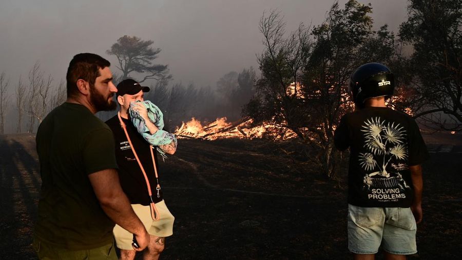 Penduduk setempat berdiri di samping mobil saat asap mengepul dari kebakaran hutan yang terjadi di desa Varnavas, dekat Athena, Yunani, 11 Agustus 2024. (REUTERS/Hilary Swift)