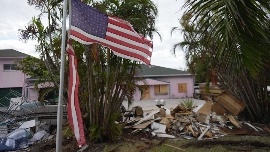 A tattered American flag flaps outside a home as furniture and household items damaged by Hurricane Helene flooding sit piled along the street awaiting pickup, ahead of the arrival of Hurricane Milton, in Holmes Beach on Anna Maria Island, Fla., Tuesday, Oct. 8, 2024. (AP Photo/Rebecca Blackwell)
