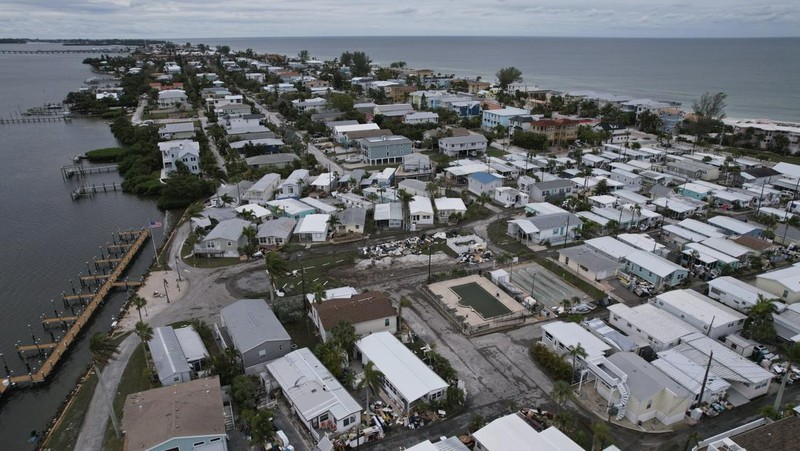 A tattered American flag flaps outside a home as furniture and household items damaged by Hurricane Helene flooding sit piled along the street awaiting pickup, ahead of the arrival of Hurricane Milton, in Holmes Beach on Anna Maria Island, Fla., Tuesday, Oct. 8, 2024. (AP Photo/Rebecca Blackwell)