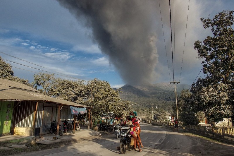Warga berkendara dengan latar belakang Gunung Lewotobi Laki Laki memuntahkan material vulkanik saat terjadi letusan di Flores Timur, Indonesia, Sabtu, 9 November 2024.(Photo by ARNOLD WELIANTO / AFP)