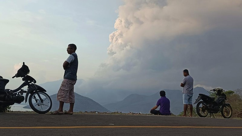 Warga berkendara dengan latar belakang Gunung Lewotobi Laki Laki memuntahkan material vulkanik saat terjadi letusan di Flores Timur, Indonesia, Sabtu, 9 November 2024.(Photo by ARNOLD WELIANTO / AFP)