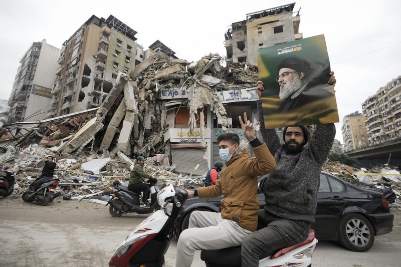 Men carry Hezbollah flags and a picture depicting late Hezbollah leader Sayyed Hassan Nasrallah, as they drive past damaged buildings at the entrance of Beirut's southern suburbs, after a ceasefire between Israel and Hezbollah took effect, Lebanon November 27, 2024. REUTERS/Mohamed Azakir     TPX IMAGES OF THE DAY