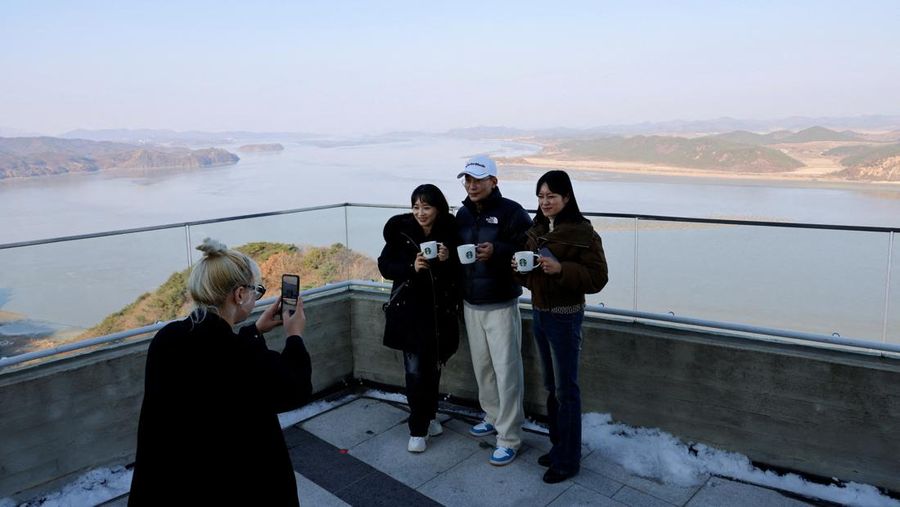 Pelanggan yang memegang secangkir kopi berpose untuk berfoto dengan desa propaganda Korea Utara Kaepoong di latar belakang, di puncak Observatorium Puncak Aegibong, selatan zona demiliterisasi (DMZ), yang memisahkan kedua Korea, di Gimpo, Korea Selatan, 29 November 2024. (REUTERS/Kim Soo-hyeon)