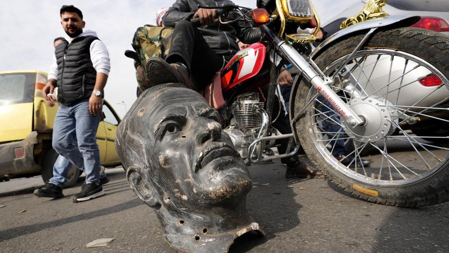 Orang-orang berdiri di dekat patung mantan presiden Suriah Hafez al-Assad yang rusak setelah pemberontak Suriah mengumumkan bahwa mereka telah menggulingkan Presiden Bashar al-Assad, di Qamishli, Suriah, 8 Desember 2024. (REUTERS/Orhan Qereman)