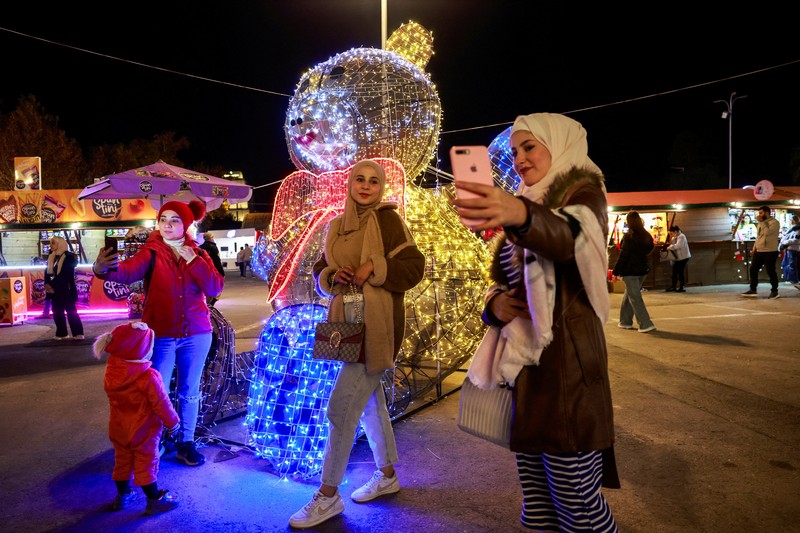 Para wanita memegang bendera yang diadopsi oleh penguasa baru Suriah, saat mereka berpose untuk difoto di dekat dekorasi Natal di Damaskus, setelah Bashar al-Assad digulingkan, di Damaskus, Suriah, 19 Desember 2024. (REUTERS/Ammar Awad)