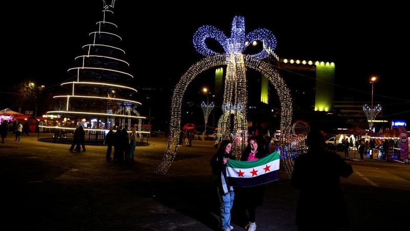 Para wanita memegang bendera yang diadopsi oleh penguasa baru Suriah, saat mereka berpose untuk difoto di dekat dekorasi Natal di Damaskus, setelah Bashar al-Assad digulingkan, di Damaskus, Suriah, 19 Desember 2024. (REUTERS/Ammar Awad)