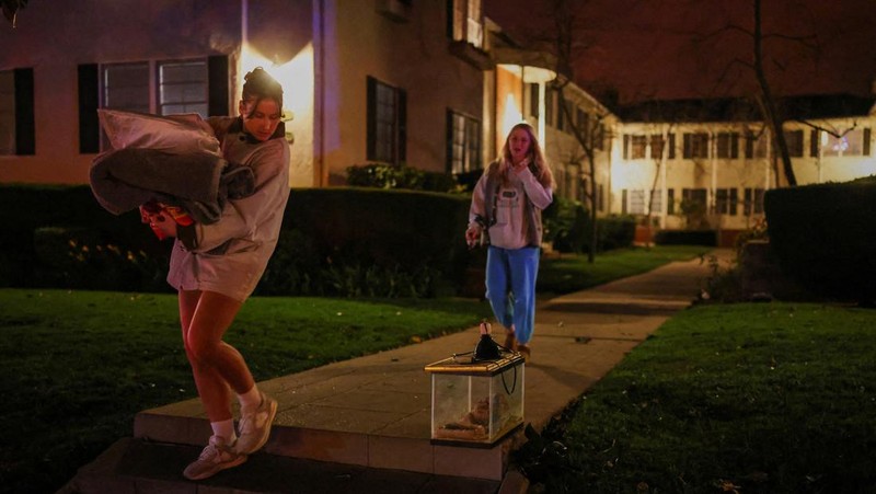 Seorang yang membawa bendera berjalan menjauh dari rumah yang terbakar saat angin kencang yang memicu kebakaran hutan yang dahsyat di wilayah Los Angeles memaksa orang-orang untuk mengungsi, di Kebakaran Eaton di Altadena, California, AS, 8 Januari 2025. (REUTERS/David Swanson)