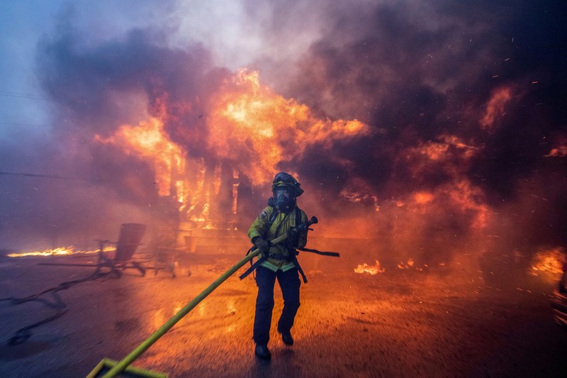 Seorang yang membawa bendera berjalan menjauh dari rumah yang terbakar saat angin kencang yang memicu kebakaran hutan yang dahsyat di wilayah Los Angeles memaksa orang-orang untuk mengungsi, di Kebakaran Eaton di Altadena, California, AS, 8 Januari 2025. (REUTERS/David Swanson)