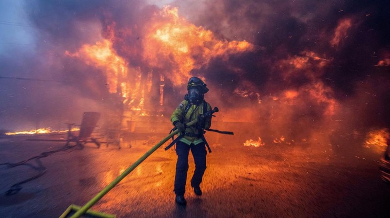 Seorang yang membawa bendera berjalan menjauh dari rumah yang terbakar saat angin kencang yang memicu kebakaran hutan yang dahsyat di wilayah Los Angeles memaksa orang-orang untuk mengungsi, di Kebakaran Eaton di Altadena, California, AS, 8 Januari 2025. (REUTERS/David Swanson)