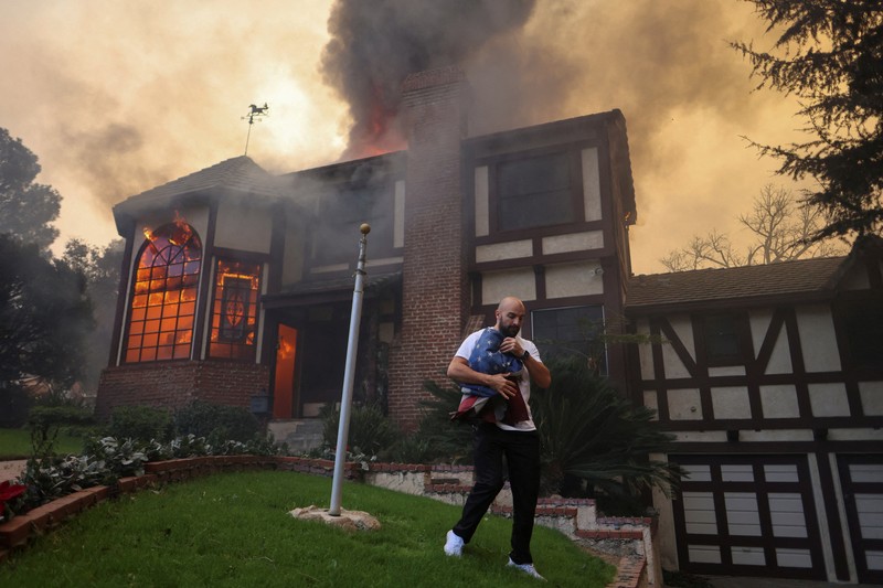 Seorang yang membawa bendera berjalan menjauh dari rumah yang terbakar saat angin kencang yang memicu kebakaran hutan yang dahsyat di wilayah Los Angeles memaksa orang-orang untuk mengungsi, di Kebakaran Eaton di Altadena, California, AS, 8 Januari 2025. (REUTERS/David Swanson)