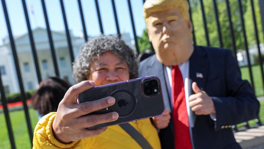A tourist uses an Apple iPhone to take a selfie with Donald Trump impersonator Ed Weiskopf in front of the White House in Washington, D.C., U.S., April 9, 2025. REUTERS/Jonathan Ernst