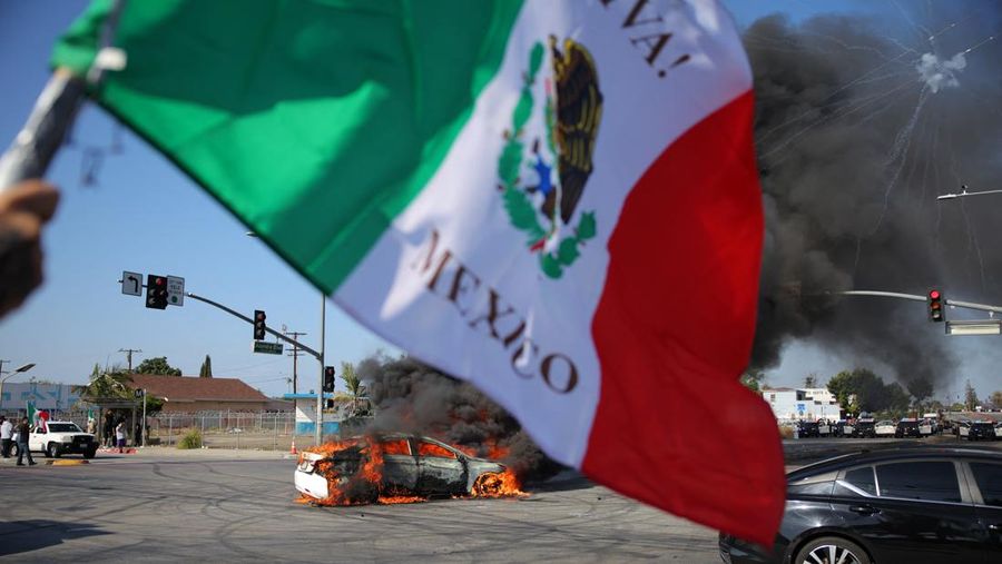 Seorang pria melambaikan bendera Meksiko di dekat mobil yang terbakar setelah beberapa kali ditahan oleh Imigrasi dan Bea Cukai (ICE), di kota Paramount, Los Angeles County, California, AS, 7 Juni 2025. REUTERS/Daniel Cole