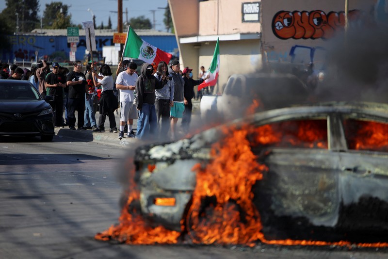 A Mexican flag waves as a car burns in flames following multiple detentions by Immigration and Customs Enforcement (ICE), in the Los Angeles County city of Paramount, California, U.S., June 7, 2025.   REUTERS/Daniel Cole