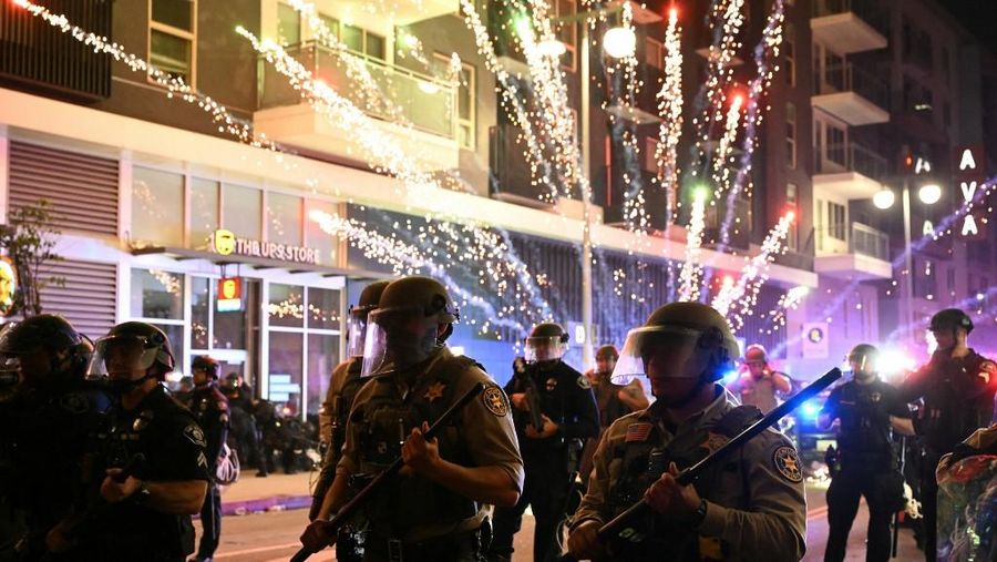 Kembang api meledak di belakang deputi departemen Sheriff Ventura County yang mengenakan perlengkapan anti huru hara selama protes sebagai tanggapan terhadap operasi imigrasi federal di lingkungan Little Tokyo di pusat kota Los Angeles, pada 9 Juni 2025. (AFP/RONALDO SCHEMIDT)