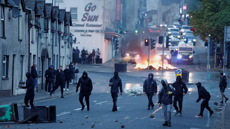 Orang-orang berjalan saat api membubung pada malam kedua kerusuhan, di Ballymena, Irlandia Utara, 10 Juni 2025. (REUTERS/Clodagh Kilcoyne)