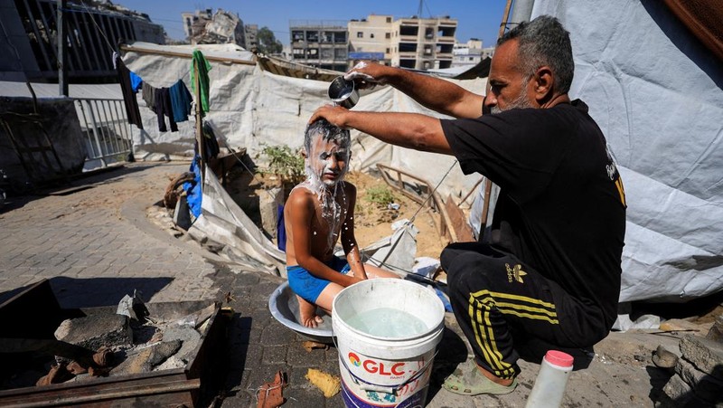 Seorang pria Palestina, yang mengungsi akibat serangan Israel, memandikan putranya di dalam tempat penampungan mereka di tengah teriknya musim panas, di Kota Gaza, 3 Juli 2025. (REUTERS/Dawoud Abu Alkas)