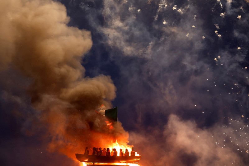 Tumpukan api unggun loyalis terlihat bersama model perahu berisi manekin yang digambarkan sebagai migran berjaket pelampung dengan tanda bertuliskan 