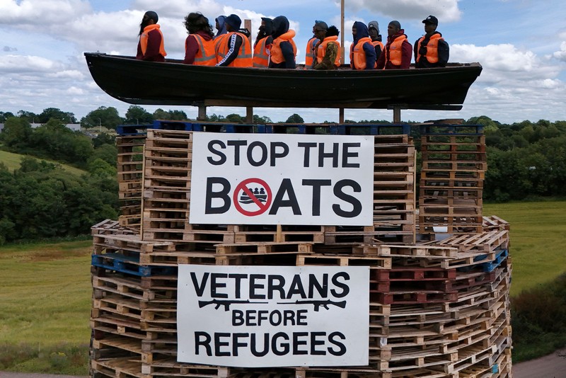 Tumpukan api unggun loyalis terlihat bersama model perahu berisi manekin yang digambarkan sebagai migran berjaket pelampung dengan tanda bertuliskan Tumpukan api unggun loyalis terlihat bersama model perahu berisi manekin yang digambarkan sebagai migran berjaket pelampung dengan tanda bertuliskan