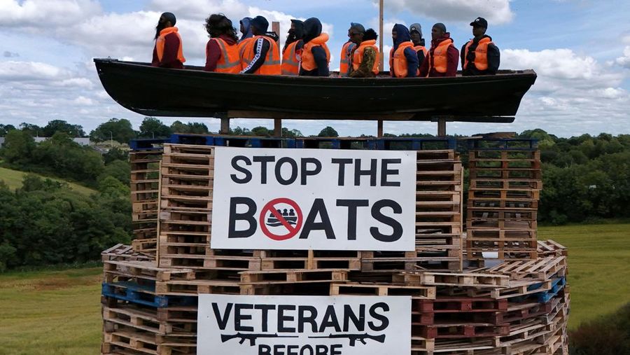 Tumpukan api unggun loyalis terlihat bersama model perahu berisi manekin yang digambarkan sebagai migran berjaket pelampung dengan tanda bertuliskan 