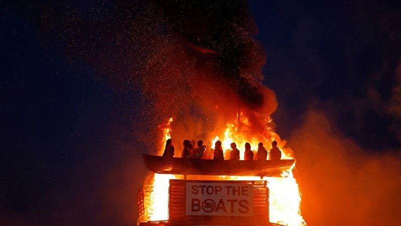 A loyalist bonfire burns after being set alight, featuring a model effigy of a boat containing mannequins portrayed as migrants in lifejackets, with a sign reading 