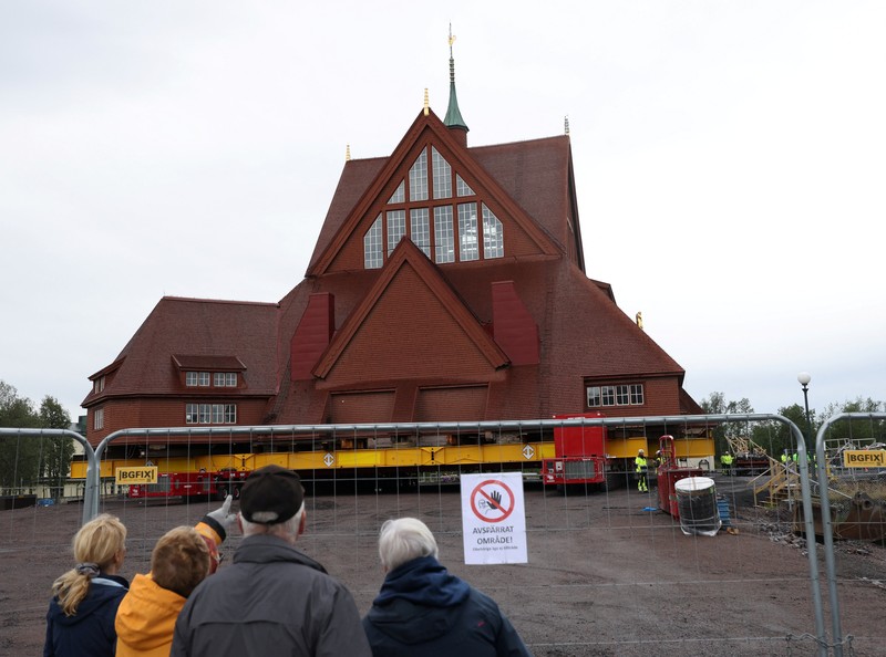 Orang-orang menggunakan ponsel mereka saat gereja kayu tua Kiruna dipindahkan selama perjalanan relokasi dua hari ke lokasi baru di sebelah pemakaman, untuk menyelamatkan dinding kayunya dari penurunan tanah dan perluasan tambang bijih besi bawah tanah terbesar di dunia, di Kiruna, Swedia 19 Agustus 2025. (REUTERS/Leonhard Foeger)|