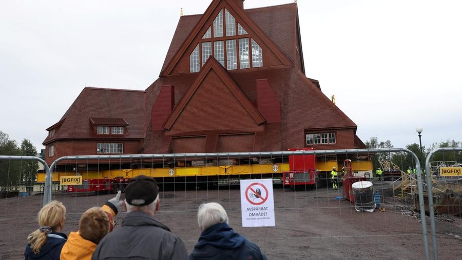 Orang-orang menggunakan ponsel mereka saat gereja kayu tua Kiruna dipindahkan selama perjalanan relokasi dua hari ke lokasi baru di sebelah pemakaman, untuk menyelamatkan dinding kayunya dari penurunan tanah dan perluasan tambang bijih besi bawah tanah terbesar di dunia, di Kiruna, Swedia 19 Agustus 2025. (REUTERS/Leonhard Foeger)|