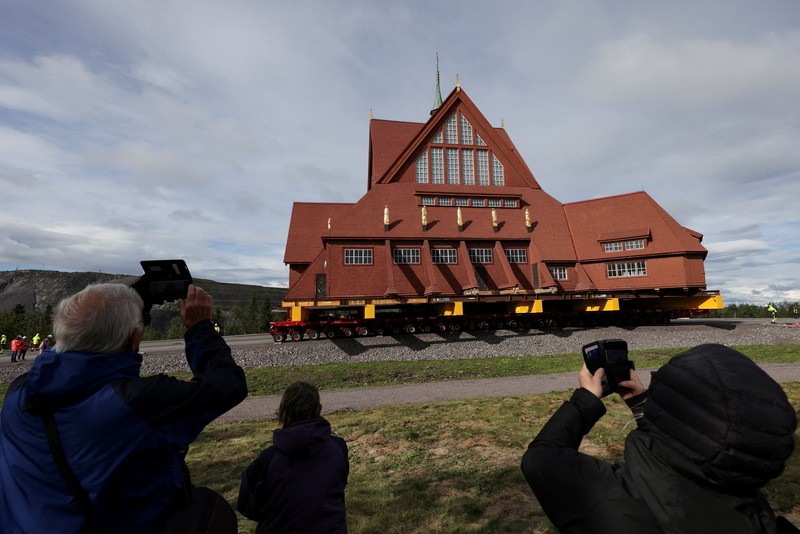 Orang-orang menggunakan ponsel mereka saat gereja kayu tua Kiruna dipindahkan selama perjalanan relokasi dua hari ke lokasi baru di sebelah pemakaman, untuk menyelamatkan dinding kayunya dari penurunan tanah dan perluasan tambang bijih besi bawah tanah terbesar di dunia, di Kiruna, Swedia 19 Agustus 2025. (REUTERS/Leonhard Foeger)|