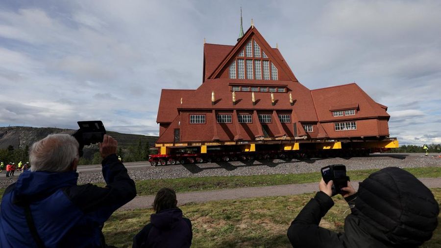 Orang-orang menggunakan ponsel mereka saat gereja kayu tua Kiruna dipindahkan selama perjalanan relokasi dua hari ke lokasi baru di sebelah pemakaman, untuk menyelamatkan dinding kayunya dari penurunan tanah dan perluasan tambang bijih besi bawah tanah terbesar di dunia, di Kiruna, Swedia 19 Agustus 2025. (REUTERS/Leonhard Foeger)|