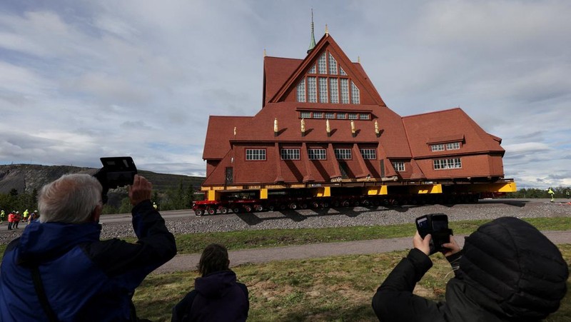 Orang-orang menggunakan ponsel mereka saat gereja kayu tua Kiruna dipindahkan selama perjalanan relokasi dua hari ke lokasi baru di sebelah pemakaman, untuk menyelamatkan dinding kayunya dari penurunan tanah dan perluasan tambang bijih besi bawah tanah terbesar di dunia, di Kiruna, Swedia 19 Agustus 2025. (REUTERS/Leonhard Foeger)|