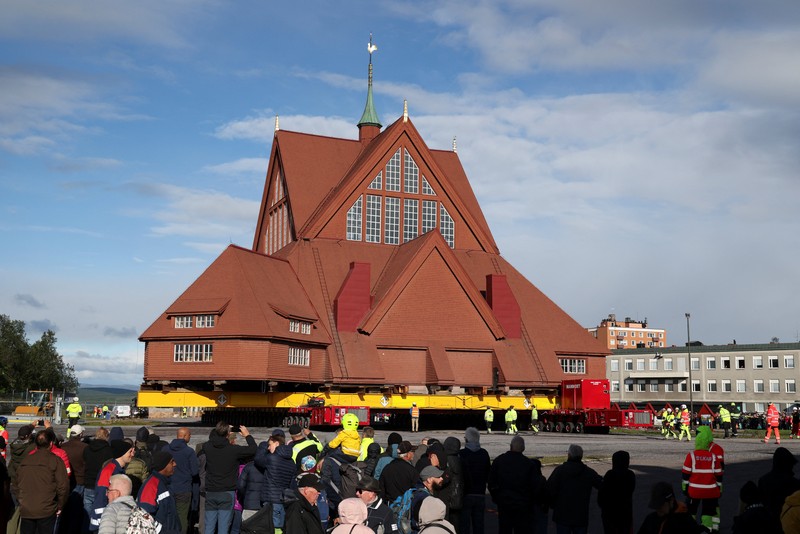 Orang-orang menggunakan ponsel mereka saat gereja kayu tua Kiruna dipindahkan selama perjalanan relokasi dua hari ke lokasi baru di sebelah pemakaman, untuk menyelamatkan dinding kayunya dari penurunan tanah dan perluasan tambang bijih besi bawah tanah terbesar di dunia, di Kiruna, Swedia 19 Agustus 2025. (REUTERS/Leonhard Foeger)|