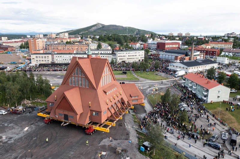 Orang-orang menggunakan ponsel mereka saat gereja kayu tua Kiruna dipindahkan selama perjalanan relokasi dua hari ke lokasi baru di sebelah pemakaman, untuk menyelamatkan dinding kayunya dari penurunan tanah dan perluasan tambang bijih besi bawah tanah terbesar di dunia, di Kiruna, Swedia 19 Agustus 2025. (REUTERS/Leonhard Foeger)|